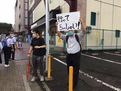 駅前では、ちょうど通勤する途中の先生方が、「行ってらっしゃい」と大きく書かれた紙を掲げて、見送りをしています。 駅前では、ちょうど通勤する途中の先生方が、「行ってらっしゃい」と大きく書かれた紙を掲げて、見送りをしています。